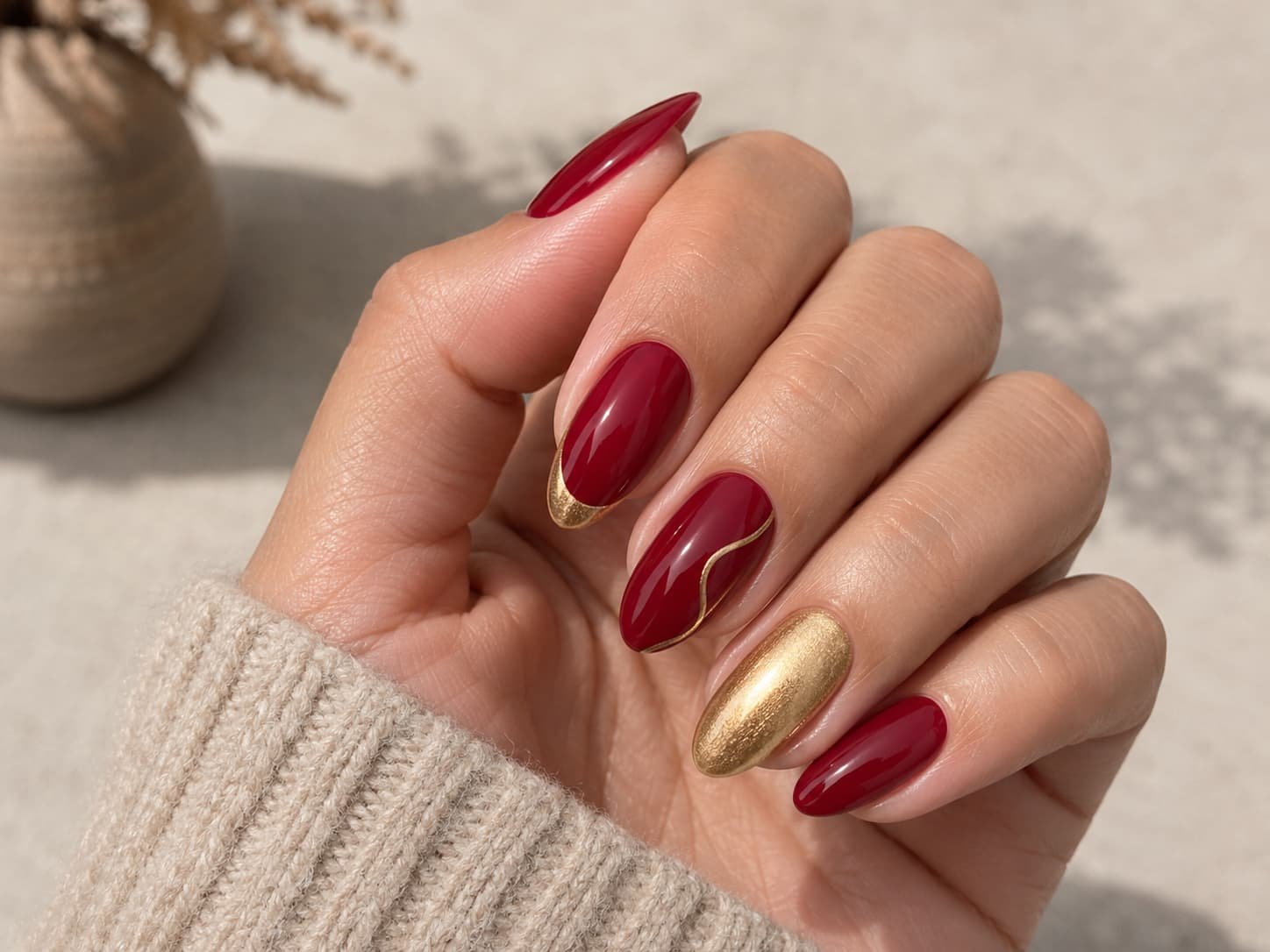 Adult hand with deep red almond nails, thin gold accents, and one metallic gold accent nail.