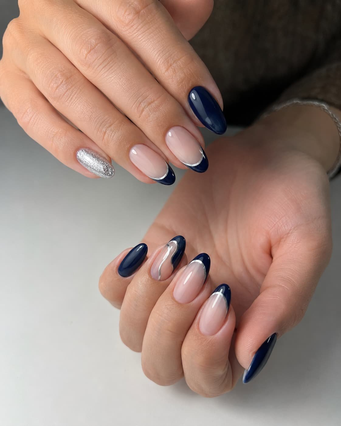 Hands with navy blue almond nails featuring glossy polish, French tips, glitter, and silver accents.